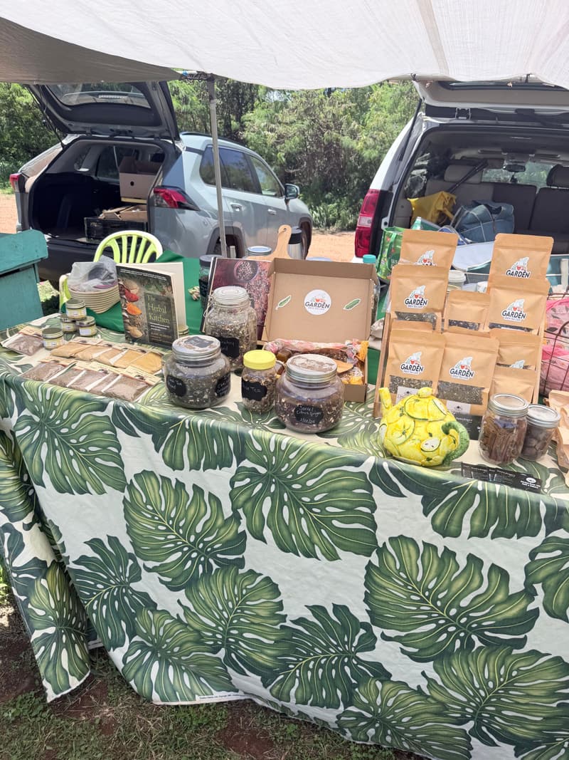 The Garden Brew booth under a market tent, with a monstera-leaf tablecloth, a yellow lemon teapot and kraft pouches of tea