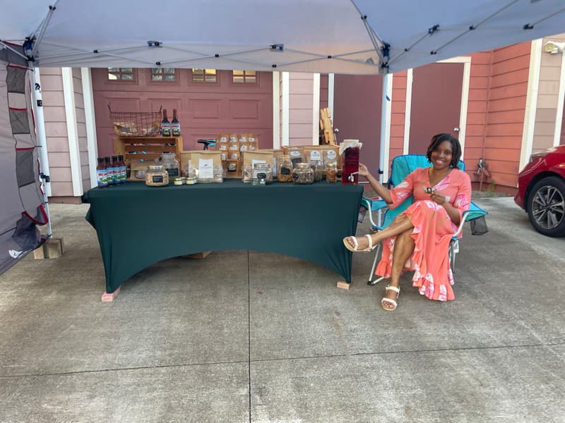 The Garden Brew Tea Cafe booth under a market tent — jars of tea on a green tablecloth, and Radia Rogers seated at the right