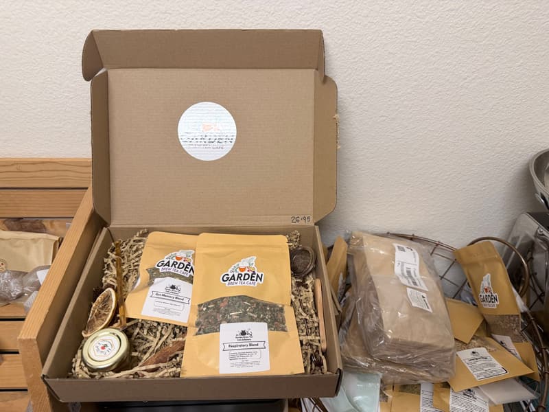 An open Garden Brew gift box lined with paper shred, holding kraft pouches of loose tea, a small jar and a dried orange slice