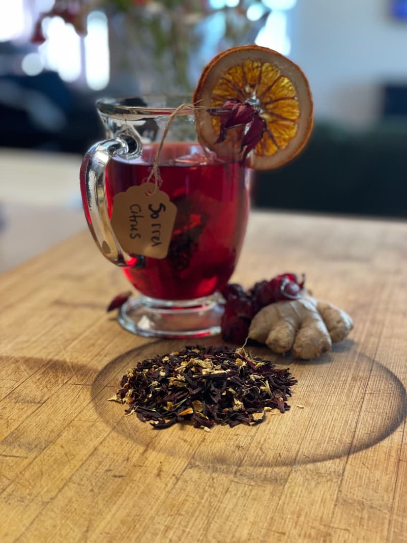 A glass mug of deep-red Sorrel Ginger Citrus tea with a dried orange slice, a knob of fresh ginger and a pile of loose blend on a wooden board