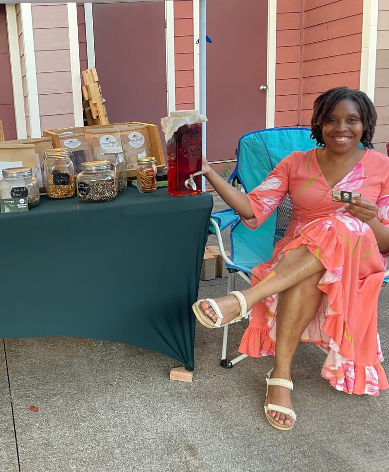 Radia Rogers, owner of Garden Brew Tea Cafe, smiling at her market booth beside a glass dispenser of red sorrel drink and jars of loose tea